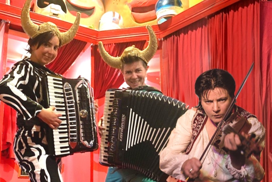 Two accordionists and a violinst wearing cow horns, performing a red-curtained tent.