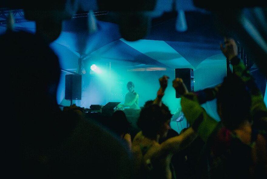 A nightclub scene with people dancing and waving their arms in the air, in the background a DJ is standing at a booth, controlling the music, large speakers on either side of them; a blue light is cast over the DJ.