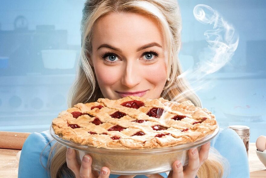A smiling woman with blond hair holding up a whole cherry pie in a dish in front of her chin, with steam rising from the pie.