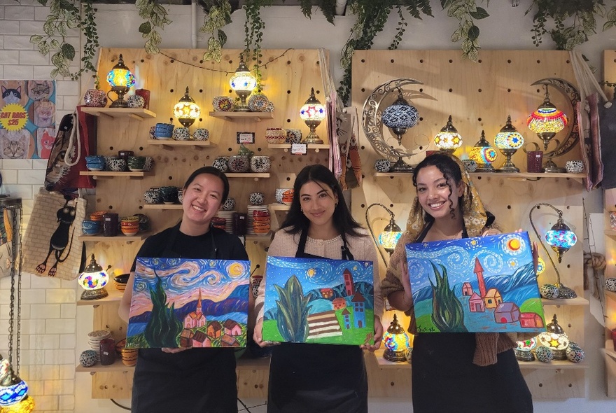 Three women each holding amateur acrylic paintings inspired by Van Gogh's Starry Night, in a workshop with pine shelving behind.