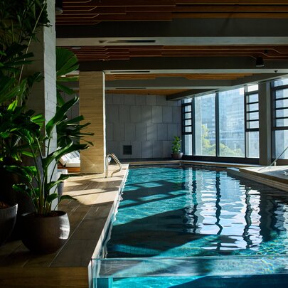 An indoor pool with green plants on one side and large windows on the other side.