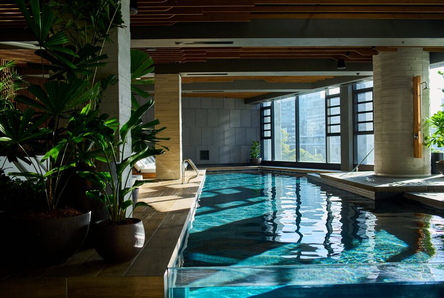 An indoor pool with green plants on one side and large windows on the other side.