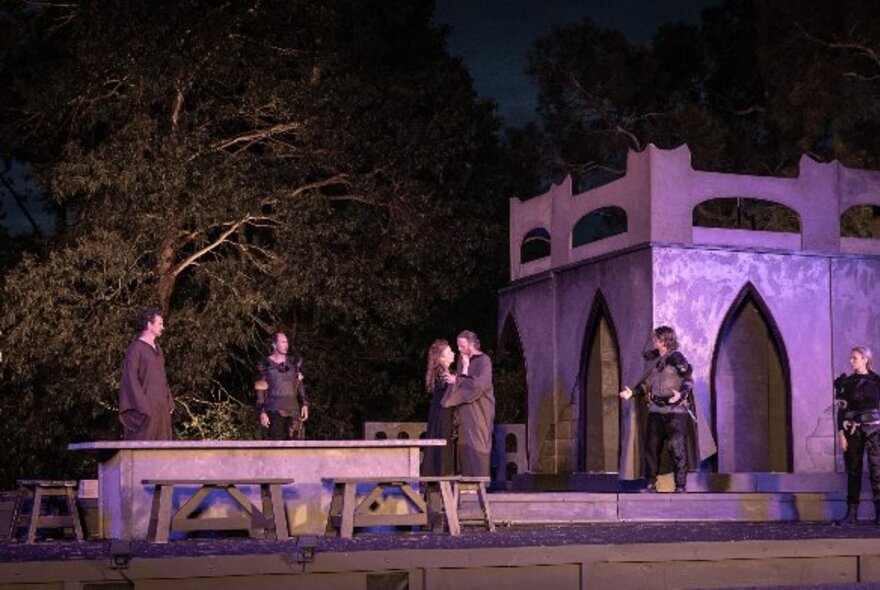 A performance of Macbeth taking place at night outdoors in the Botanic Gardens, with several actors on a stage set, two in an embrace.