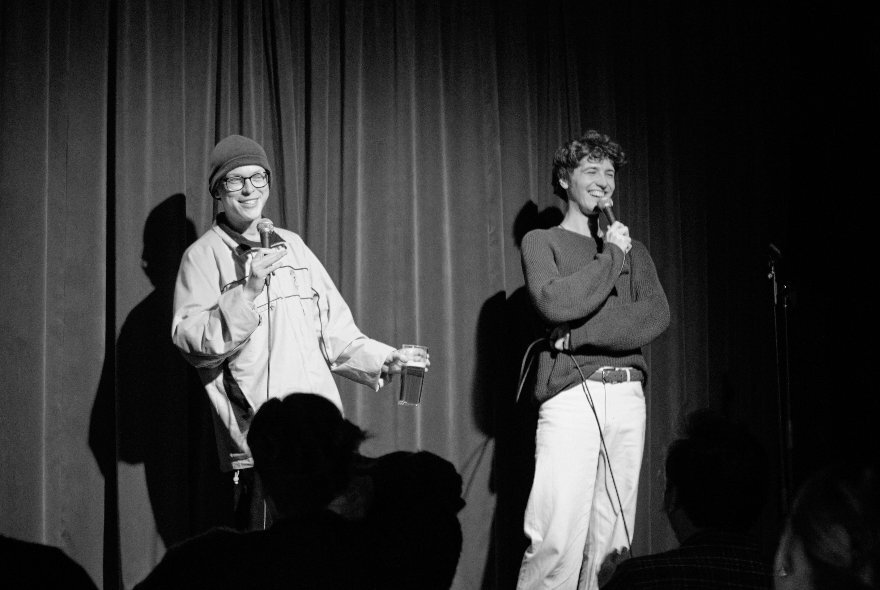 Black and white photo of two comedians on stage, both holding microphones and smiling.