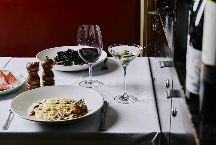 Restaurant table set for dinner with two main dishes, glass of wine, martini and condiments.