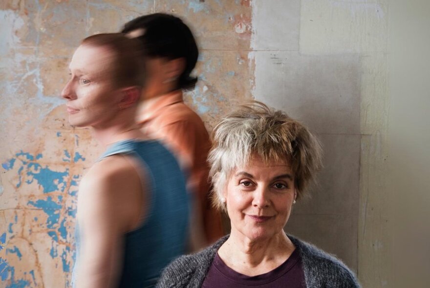 Blurred people passing an older actor with short hair, wearing grey.