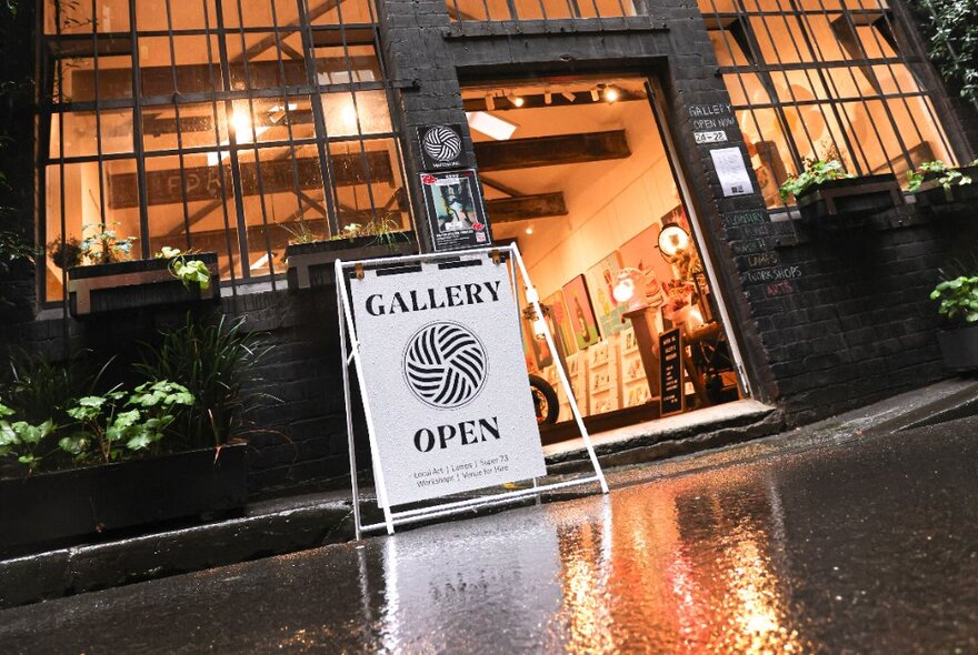 Street entrance to a studio gallery space, with a sandwich board sign out the front, and large windows; evening.