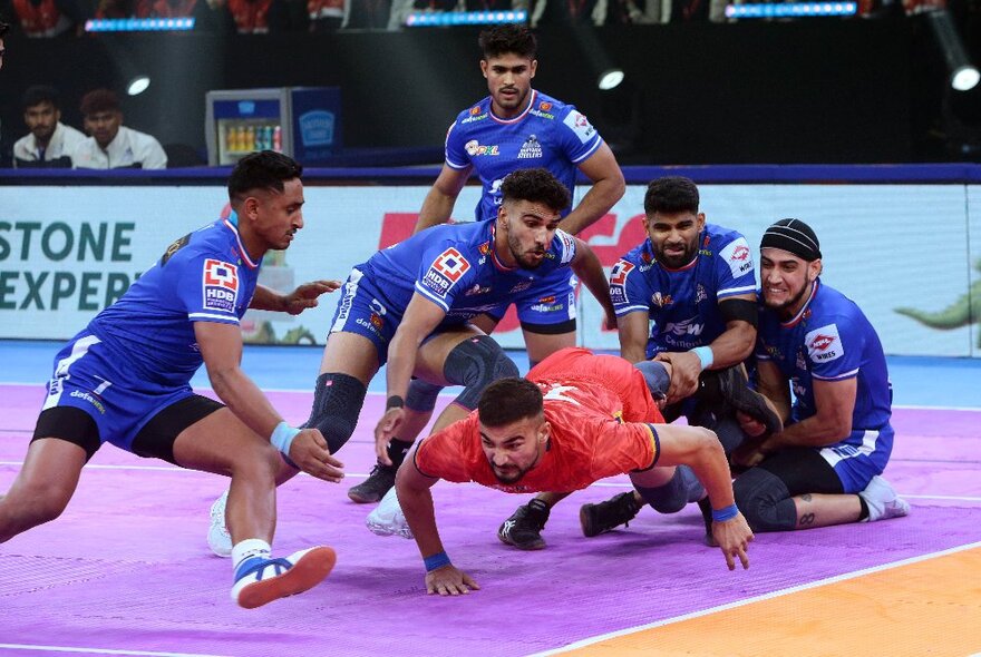 Players in opposing teams, wearing protective elbow and knee pads, running and tackling each other in the sport of kabaddi, in an indoor sporting arena.