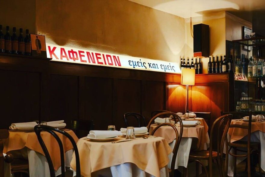 The interior of a restaurant with white linen covered tables, bentwood dining chairs and a soft neon sign bearing the restaurant's name Kafeneion that rests on a mantelpiece.