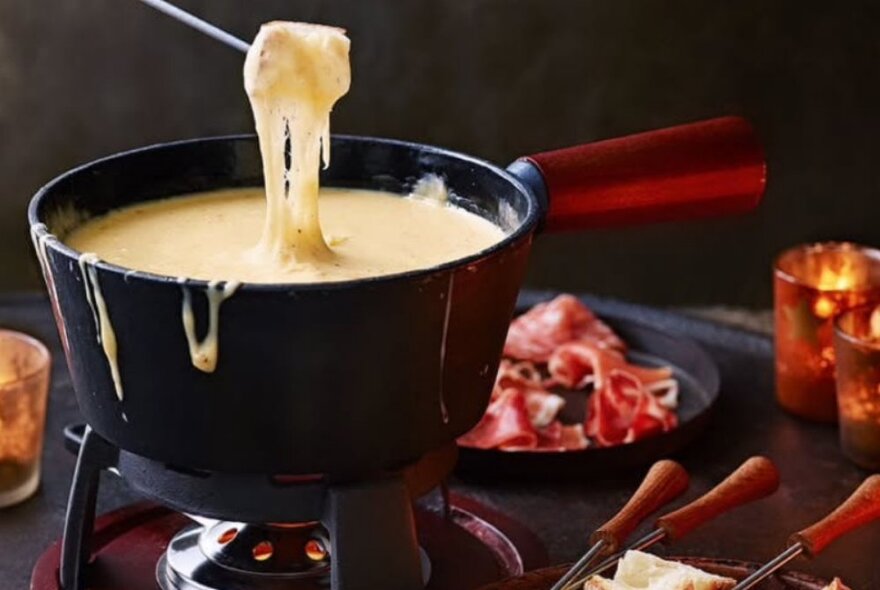 Utensil dipping into a pot of melted cheese fondue with accompaniments.