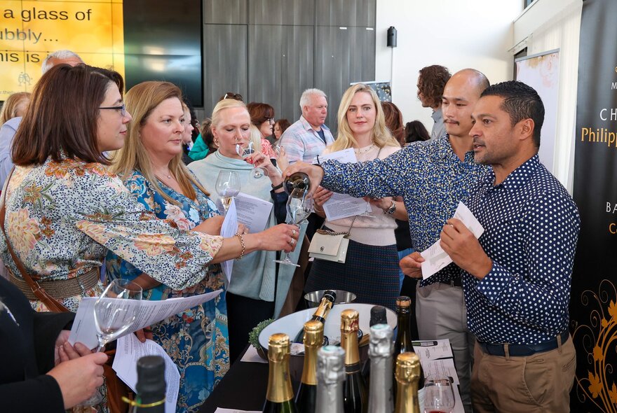 A group sampling champagne at an event, two men presenting. 