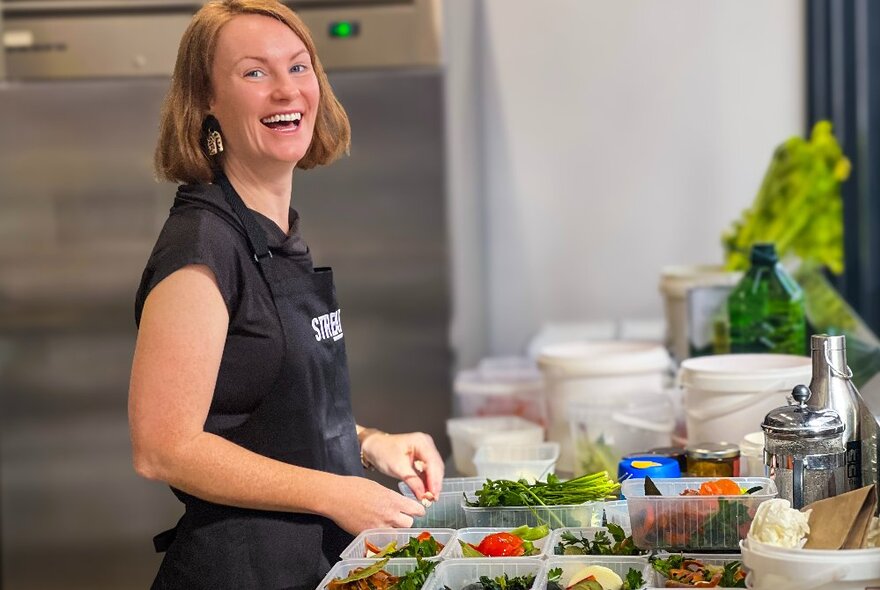 A person smiling, wearing a black apron and standing at a kitchen bench in front of containers of pre-prepared raw food.