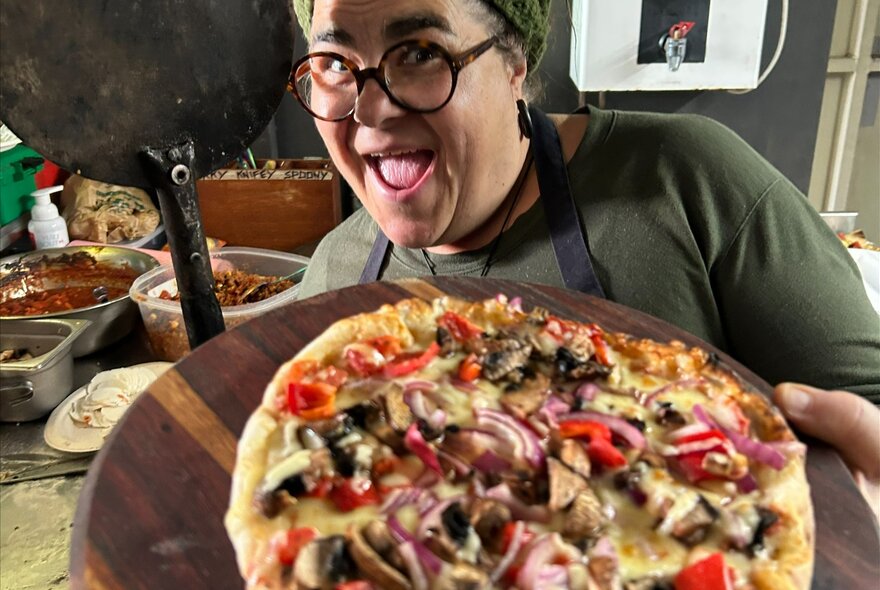 A chef or cook smiling and holding up a freshly baked wood-fired pizza in a kitchen setting.