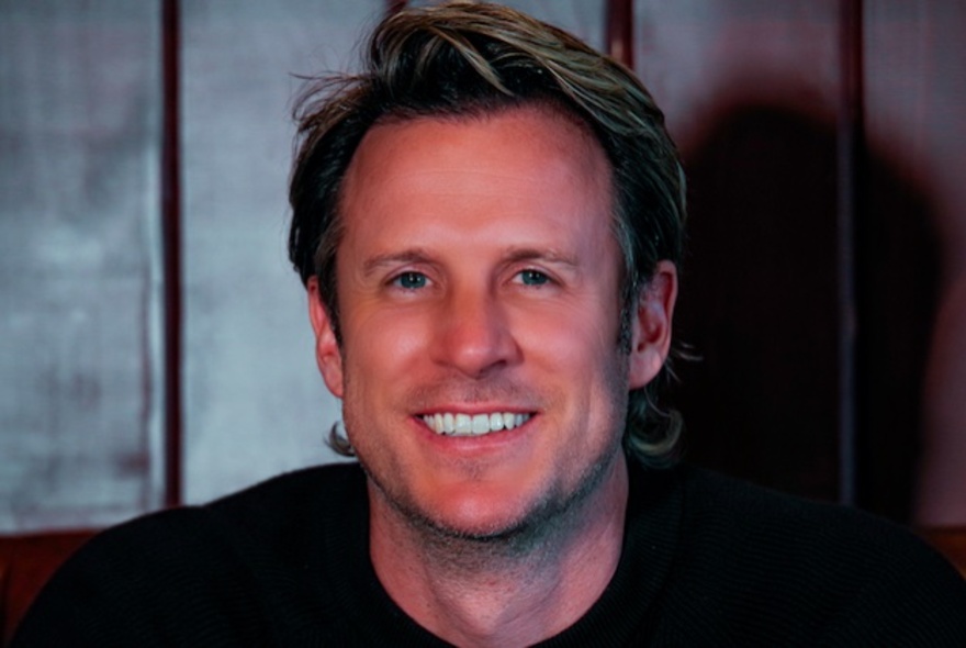 Smiling face of comedian Monty Franklin, a middle aged man wearing a dark top and posed against a panelled wall.