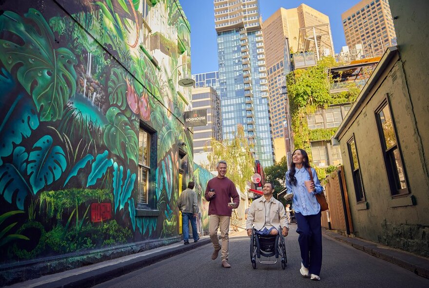 A group of friends walking past a giant jungle mural in a city laneway, one is using a wheelchair.