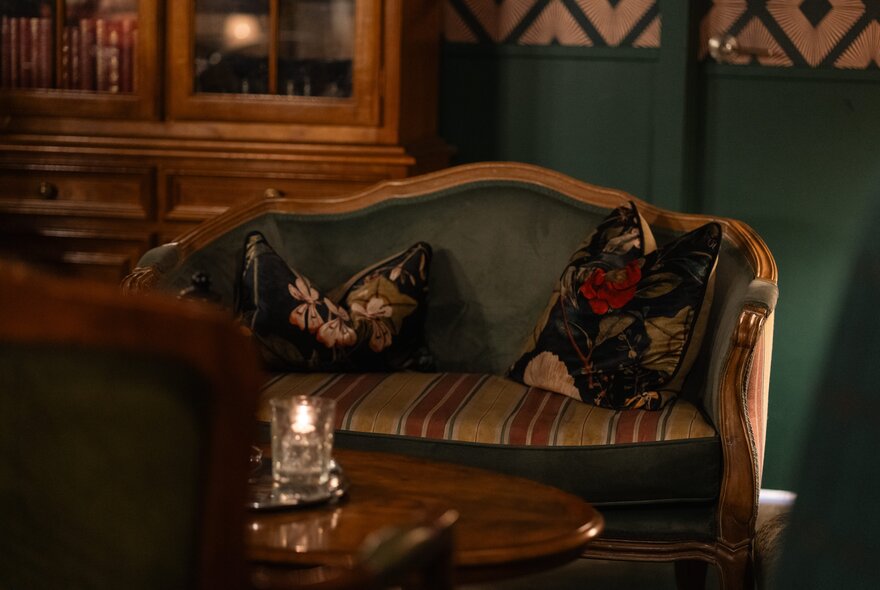 A vintage-style club featuring a classic wooden settee with green velvet cushioning and patterned throw pillows, dark green walls and a candle on a table.