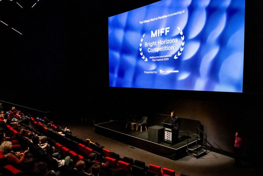 A cinema with red theatre seats and a large cinema screen showing a blue image. 