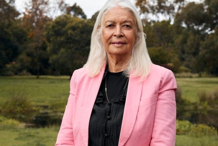 Professor Marcia Langton AO, a woman with straight white hair in her 60s, wearing a pink jacket and black top and standing in a green field.