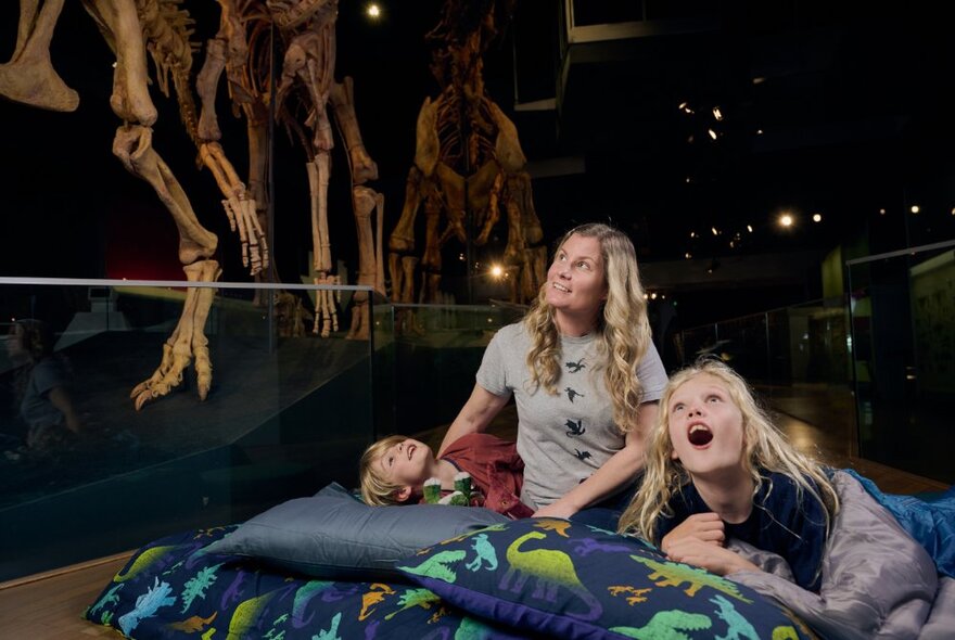A woman and two small children on a bed underneath a dinosaur skeleton at a museum sleepover