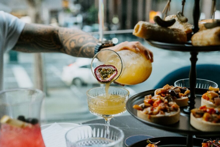 A hand pouring a drink from a carafe, with a three tiered tray of food nearby on a table.