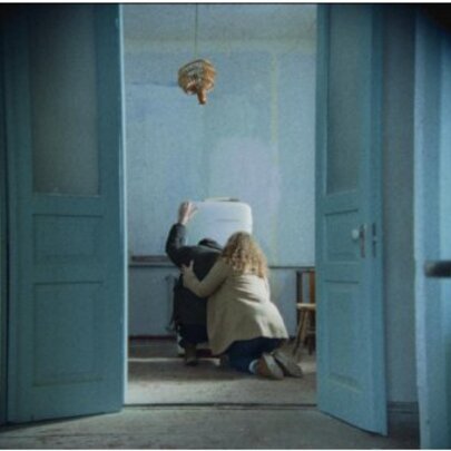 Still from a movie of a view through a doorway of a blonde woman kneeling and supporting a dark-haired individual that leans against an old fridge in a sparsely furnished room.
