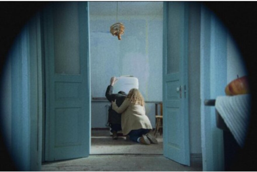Still from a movie of a view through a doorway of a blonde woman kneeling and supporting a dark-haired individual that leans against an old fridge in a sparsely furnished room.