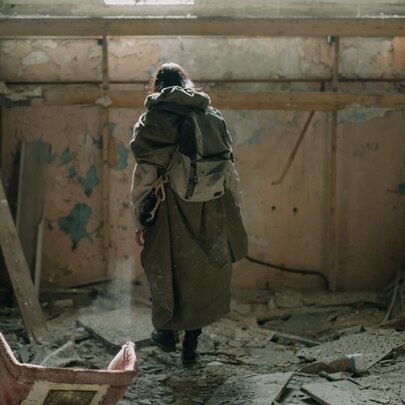 A lone figure in a long coat and backpack standing inside a ruined, debris-scattered room evoking a post-apocalyptic or dystopian atmosphere.