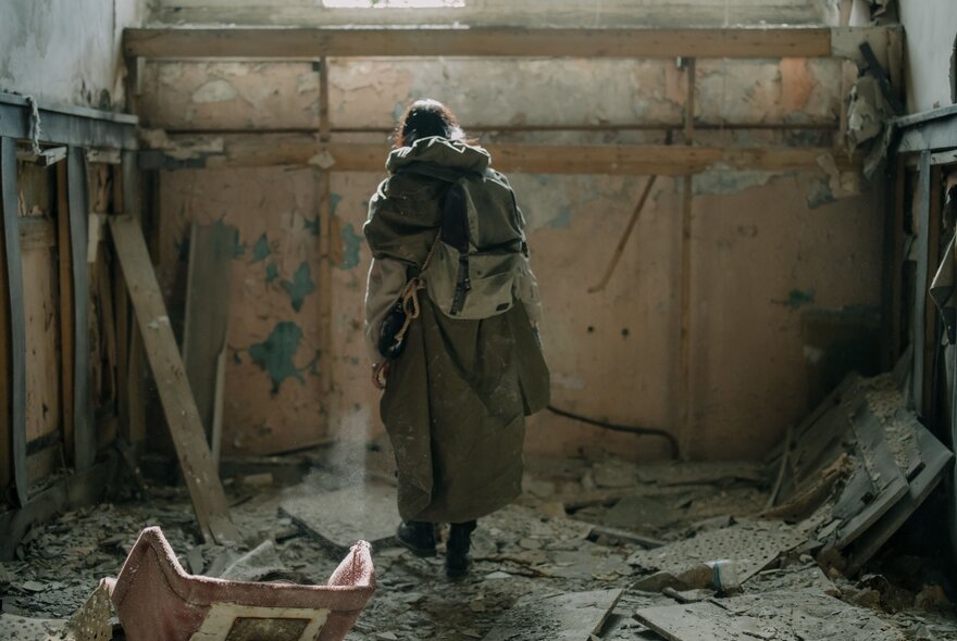 A lone figure in a long coat and backpack standing inside a ruined, debris-scattered room evoking a post-apocalyptic or dystopian atmosphere.