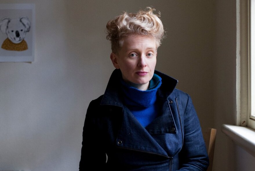 Writer Hayley Singer, wearing a navy coat, blue jumper and standing near a window inside a room.
