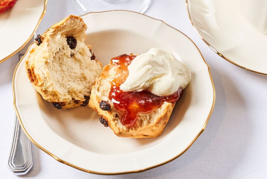 A scone, cut in half, with jam and Chantilly cream, on a gold-rimmed white side plate.