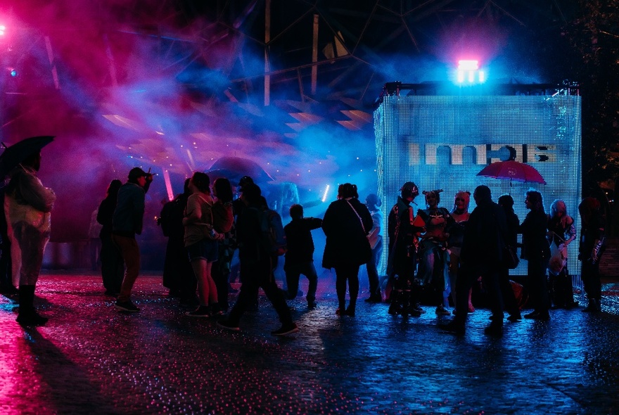 Silhouettes of people outside in the dark with pink and blue lights cast on them. 