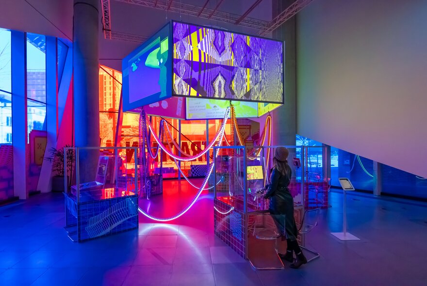 A woman standing in front of a glowing exhibition display with large screens and neon lights hanging from the ceiling.