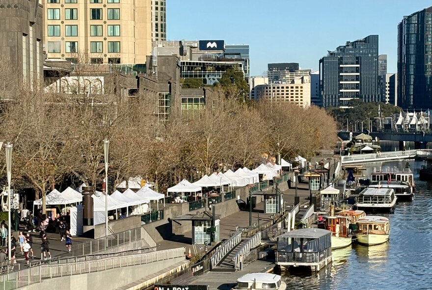 Aerial view of an outdoor market on Southbank promenade, showing the Yarra River with boats on the right, market umbrellas and stalls on the concourse and city buildings in the background.