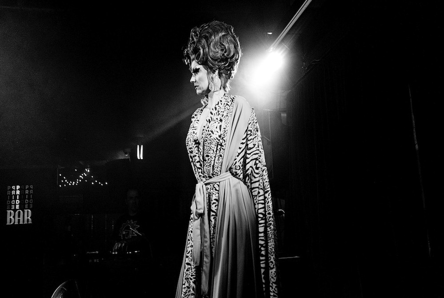 Drag performer wearing a long gown and wig under lights.