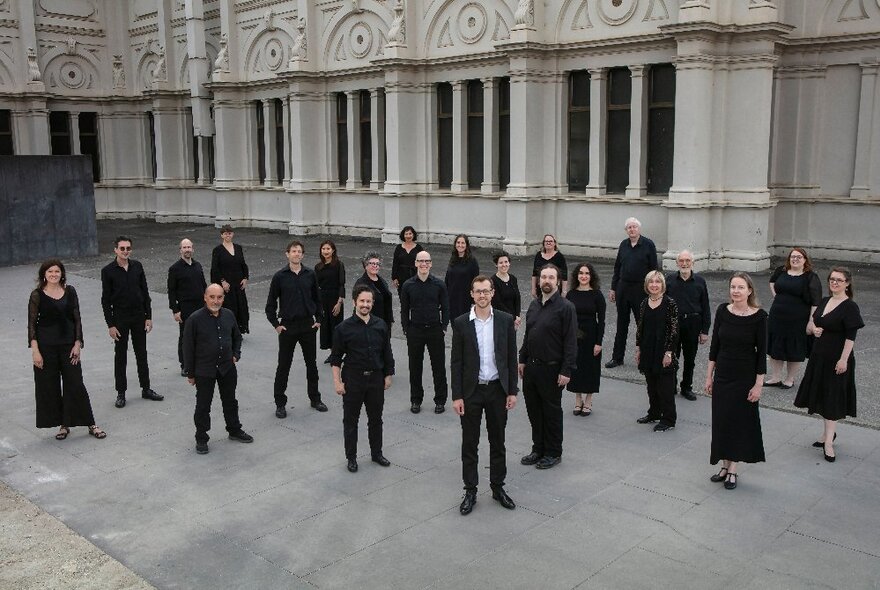 Choral group members dressed in black, standing in formation outside a classical building.