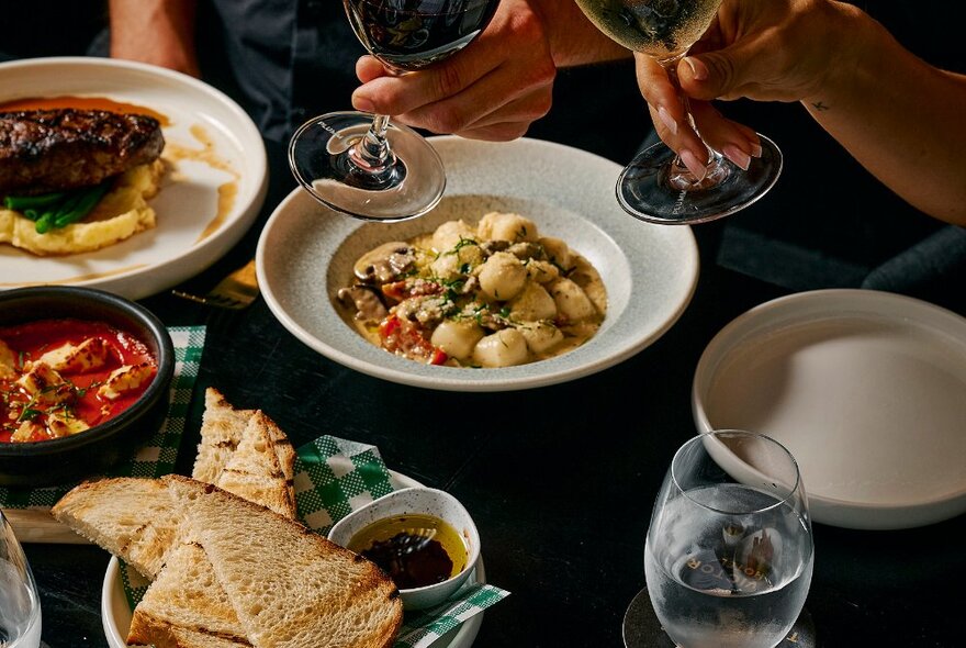 A selection of Italian food, with a gnocci dish, steak and mash and panini bread, with two hands holding wine glasses.