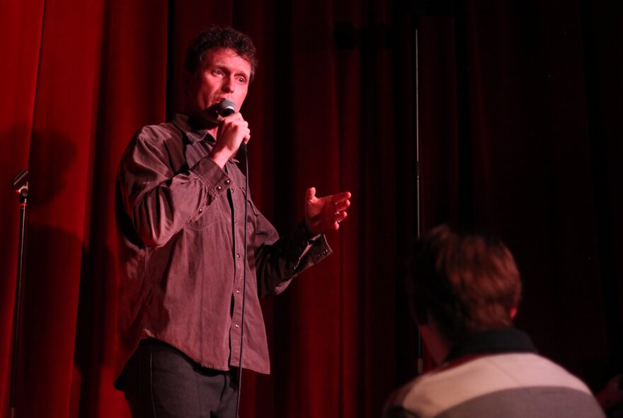 Comedian David Quirk on stage holding a microphone in his hand and performing stand up comedy.