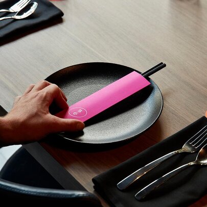 A hand places cutler in a pink paper pouch on a black dinner plate