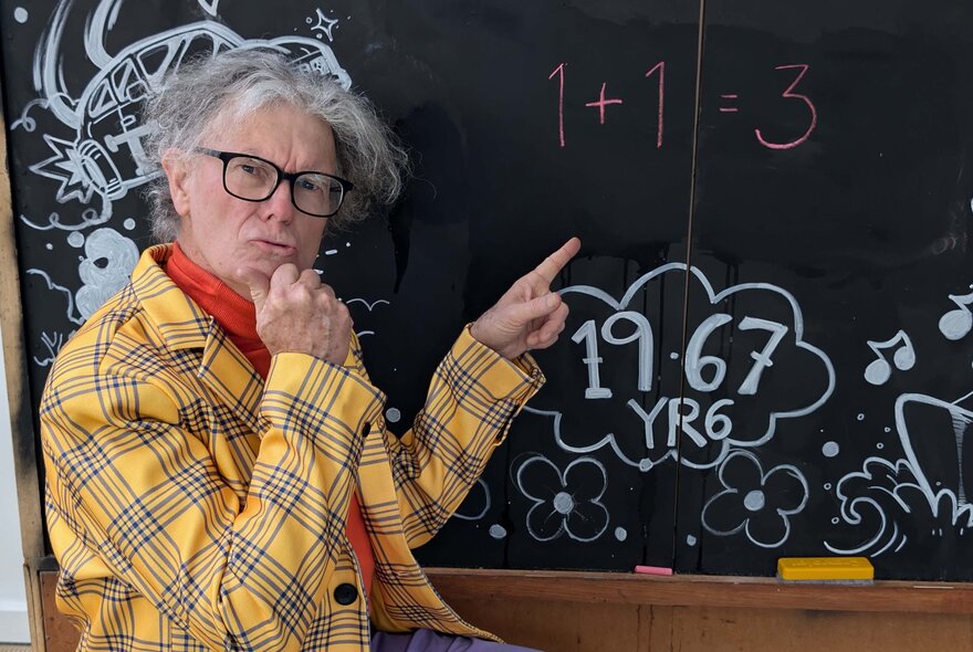 Comedian wearing yellow tartan jacket and red skivvy, with grey hair and spectacles, pointing at the blackboard, looking quizzical.