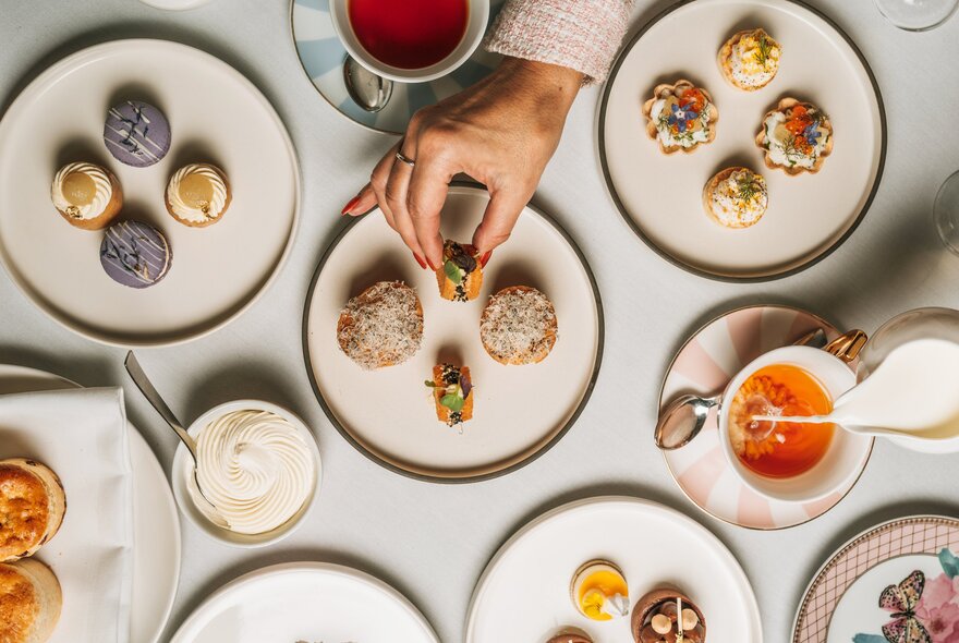 Top-down view of a table set for high tea with various small desserts on plates and a hand reaching for a treat.