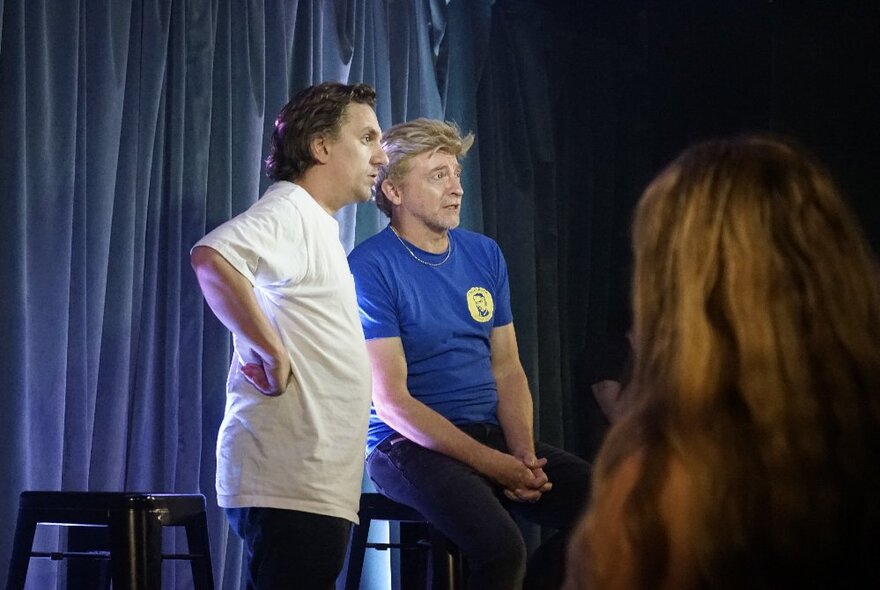 Two performers on a stage in front of a curtain, interacting with one another and improvising a scene in front of an audience.