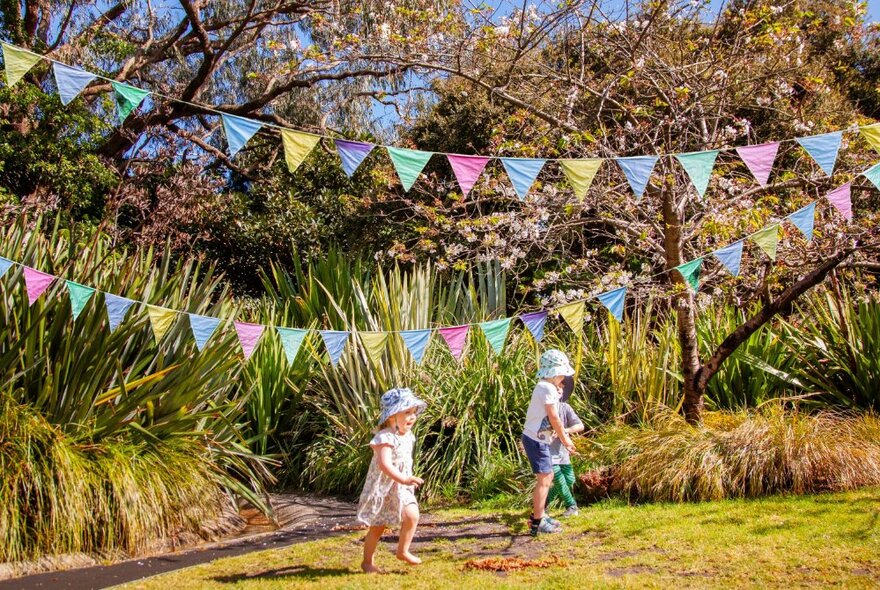 Young children running barefoot at the Ian Potter Children's Garden, with colourful bunting flying above them.