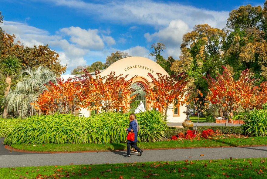 Two people walking past a conservatory in a park with orange trees on a sunny day. 