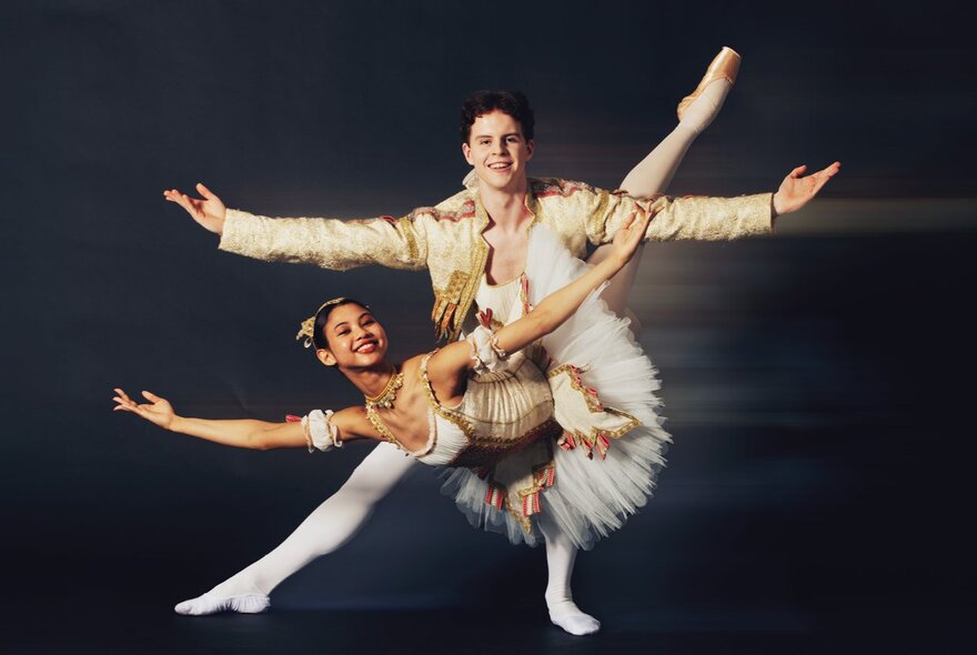 Two young adults in ballet costumes in a classical ballet position, the female wearing a white tutu with her leg raised high behind her.