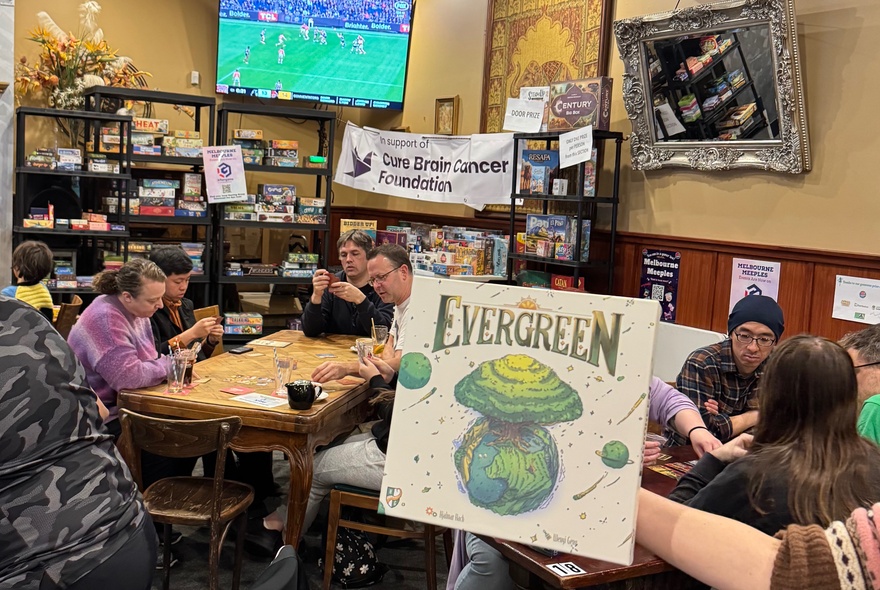 People playing boardgames at tables in a pub room with sport on a large screen.