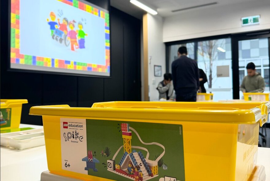 Large yellow plastic bucket in a classroom setting in front of a large screen and a group of children with teacher.