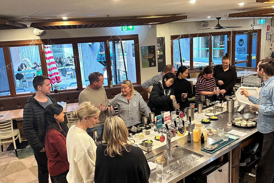A group of people standing around a stainless steel prep bench, learning to make cocktails.