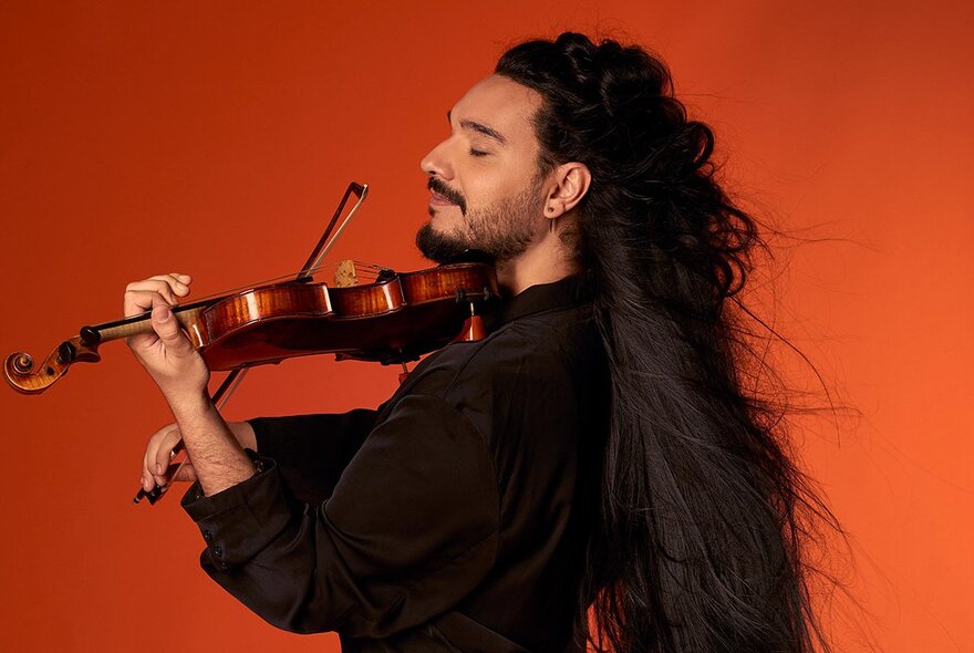 Nemanja Radulović in profile, playing his violin, his very long dark hair cascading down his back, posed against an orange background.