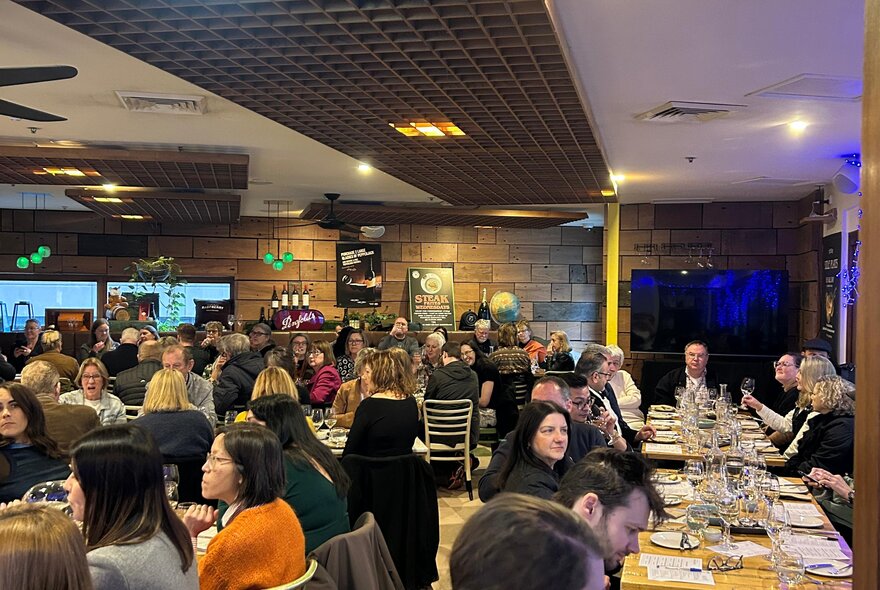 A room of people seated at tables in a restaurant, sampling wine and cheese,  listening to a person talking about wine.