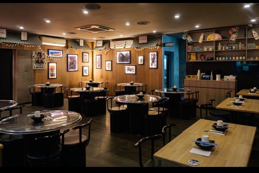 The interior of a restaurant with tables and chairs arranged in the space, small pictures hung on the wood panelled walls, and downlights. 